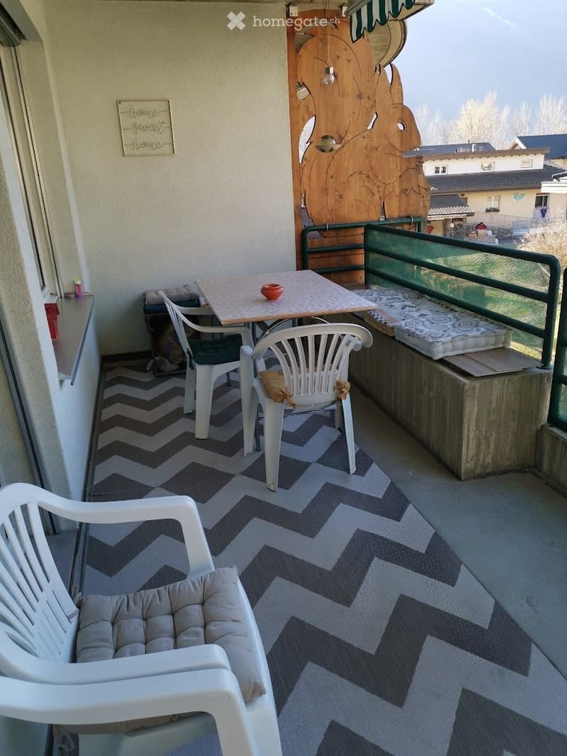 Balcony with a small table, chairs, and a cushioned seat, featuring a chevron-patterned rug and a view of the surrounding area.