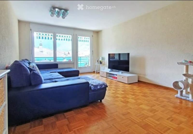 Modern living room with wooden flooring, large windows offering a view, a blue sectional sofa with an ottoman, a flat-screen TV on a white stand, and a small table near the windows.