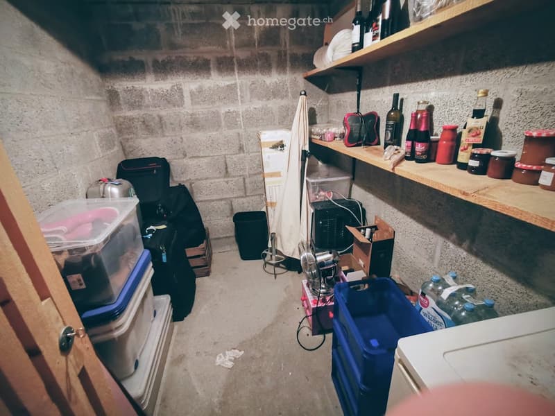 A small storage room with concrete walls and shelves holding various items, including bottles, jars, and a fan. Storage bins, backpacks, and water bottles are on the floor.