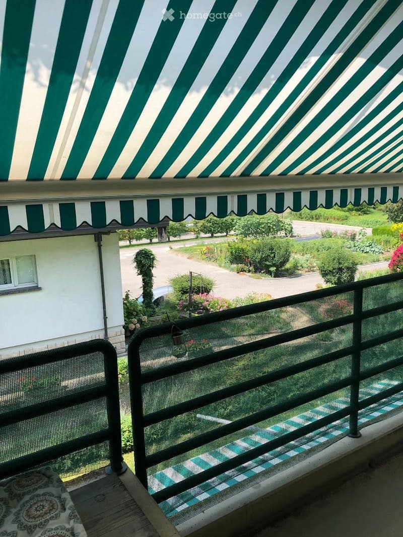 Balcony with green and white striped awning, metal railing, and view of garden and driveway.