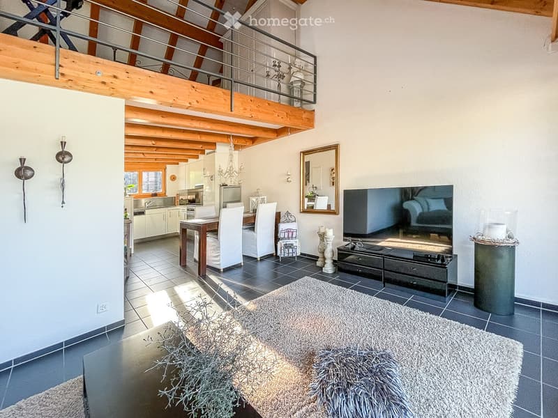 Open-plan living and dining area with exposed wooden beams, a large TV, a dining table with chairs, a sofa, and a kitchen area with modern appliances.