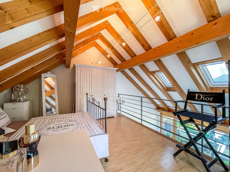 Cozy attic bedroom with wooden beams, skylights, and modern furnishings including a bed, director's chair, wardrobe, and a small table with a fan.