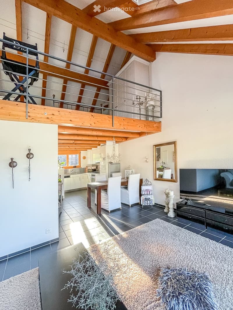 Spacious living and dining area with exposed wooden beams, modern kitchen in the background, dining table with chairs, large sofa, and decorative elements including a mirror and wall sconces.