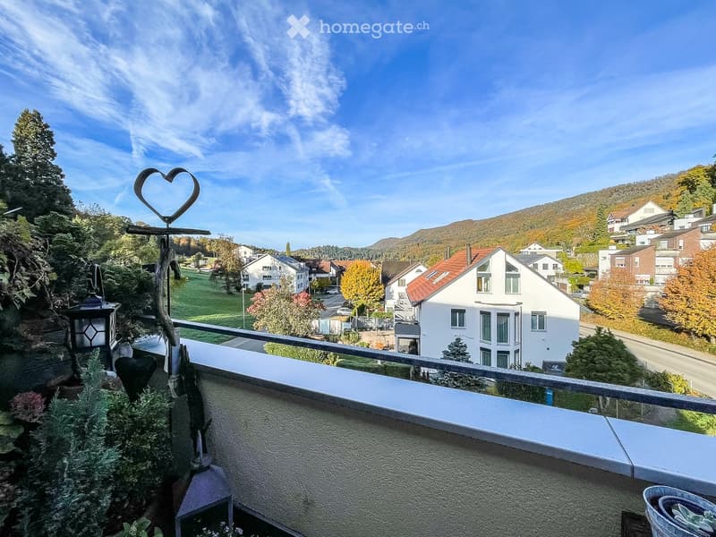 Balcony with metal railing, overlooking a residential area with houses, trees, and distant hills under a clear blue sky.