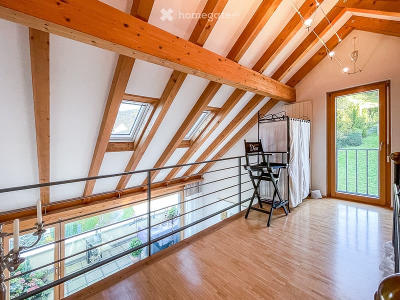 Loft-style room with wooden beams, skylights, and a balcony overlooking a green area. Features include a director's chair, candle holder, and a curtained area.