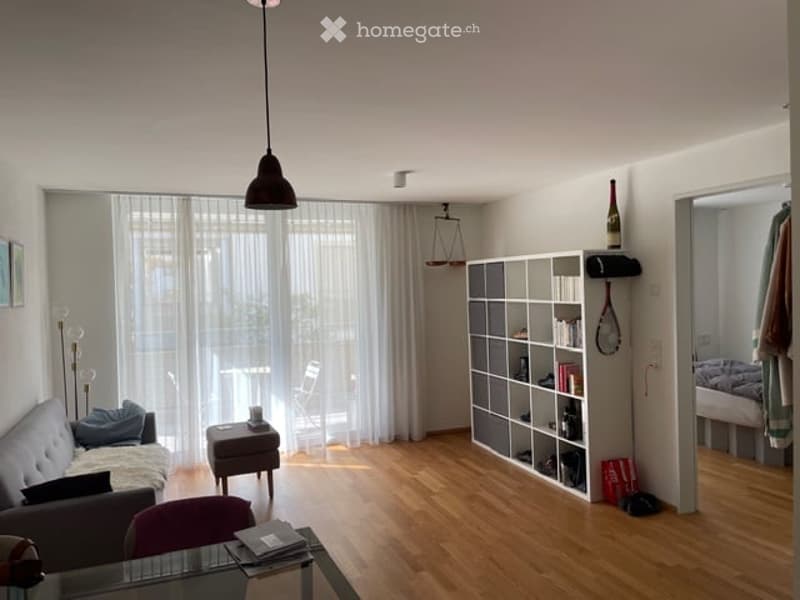 A modern, well-lit living room with wooden flooring, large windows covered by sheer curtains, a grey sofa with pillows, a small wooden stool, a white modular bookshelf, a glass coffee table, and a hanging pendant light.