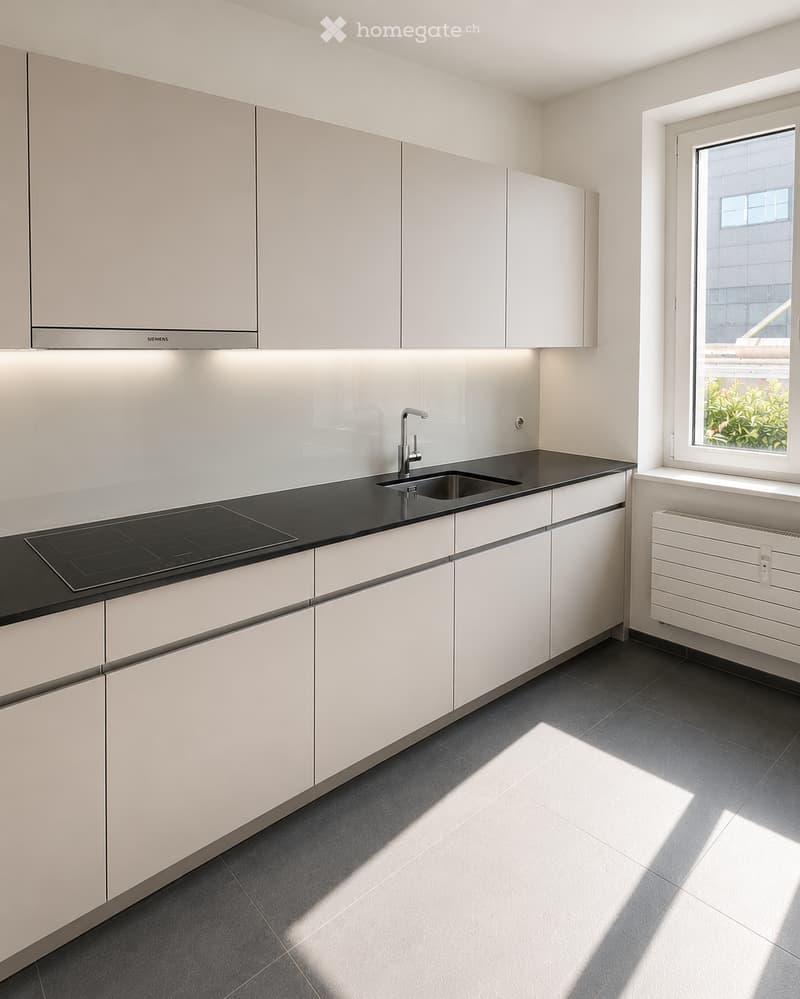 Modern kitchen with white cabinets, black countertop, induction cooktop, sink, and radiator. Natural light streams in from a window on the right.