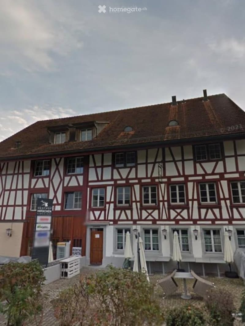 Traditional half-timbered building with red and white facade, multiple windows, and a brown tiled roof. Outdoor seating area with tables and chairs is visible in front.