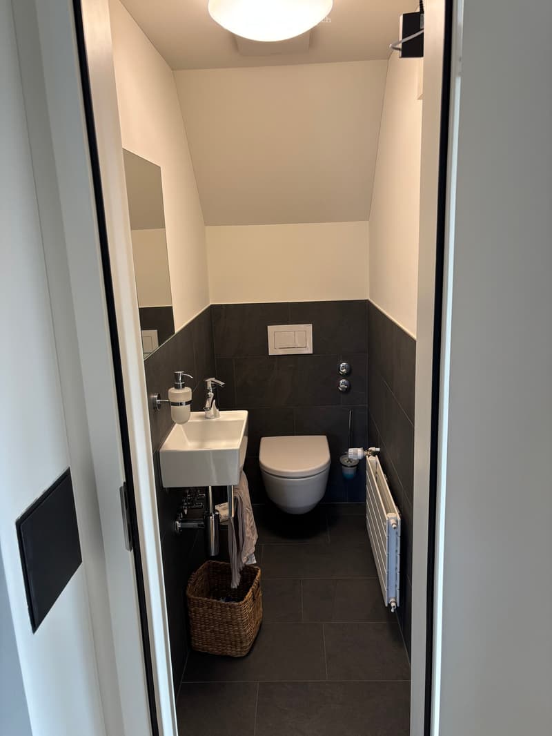Modern, compact bathroom with dark tiles, white sink, toilet, radiator, and a wicker basket on the floor.