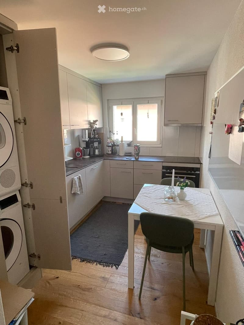 Modern kitchen with light-colored cabinets, a central island, and a small dining area. Features include a coffee maker, oven, washing machine, and a small table with chairs.