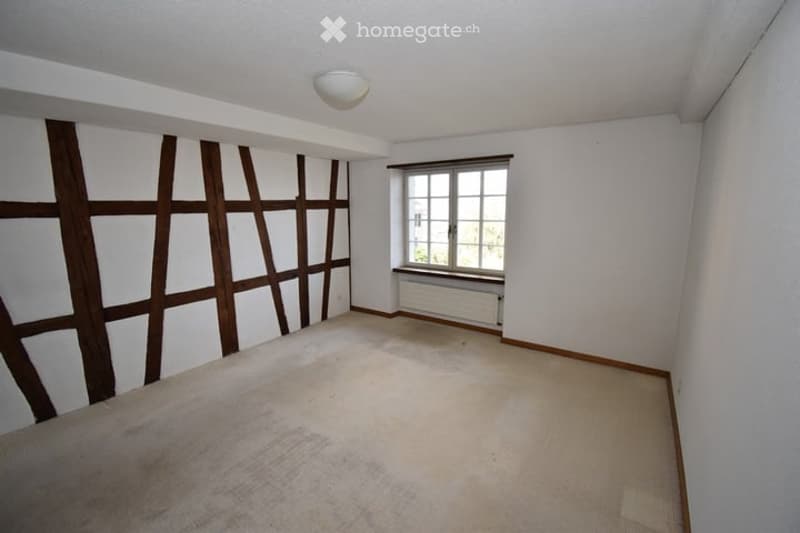 Empty room with white walls, wooden trim, carpeted floor, and a single window with radiator below.