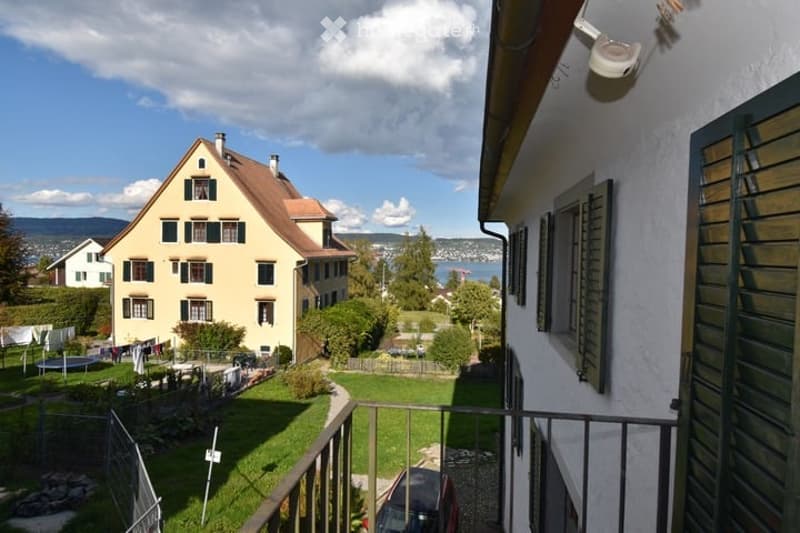 Balcony overlooking a residential area with houses, greenery, and a distant lake view under a partly cloudy sky.