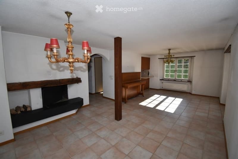 Empty room with tiled floor, white walls, fireplace with wood, large chandelier, and a wooden bench. No furniture or equipment.