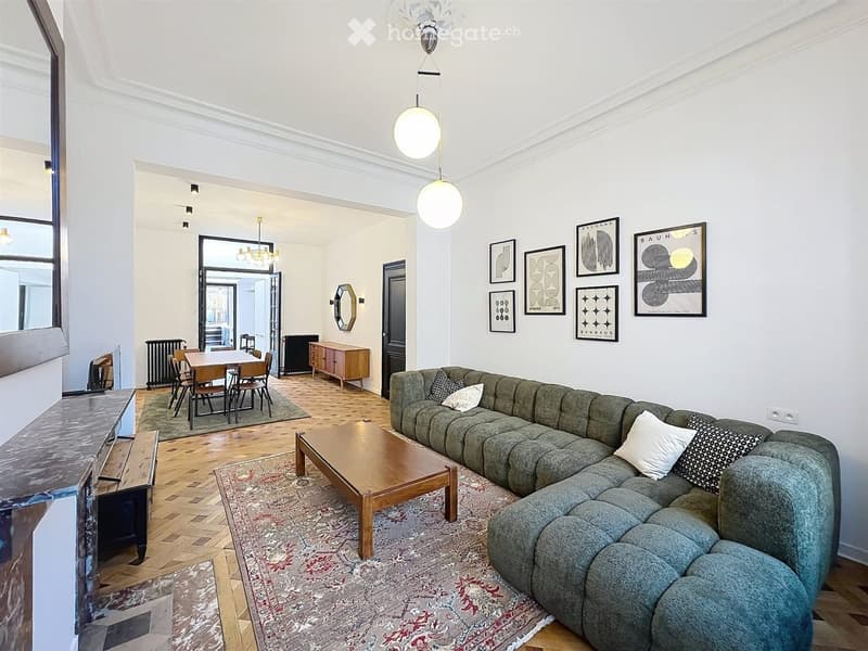Spacious living room with a grey sectional sofa, wooden coffee table, dining area with a table and chairs, and decorative artwork on the walls.