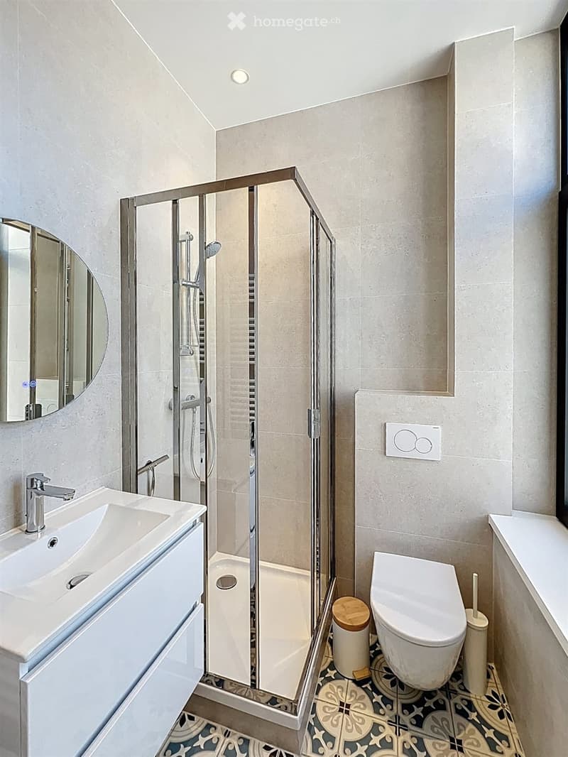 Modern bathroom with a white sink, chrome faucet, round mirror, glass-enclosed shower, toilet, and decorative tile floor.