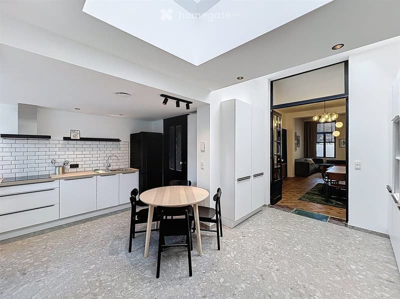 Modern kitchen with white cabinets, a round wooden dining table with four black chairs, and a sink area with a faucet. The space features a large open doorway leading to a living area with a sofa, dining table, and chandelier.