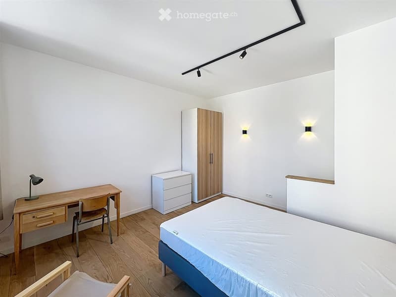 A furnished bedroom with a single bed, a wooden desk with a chair, a wardrobe, and a drawer unit. The room features wooden flooring and white walls with integrated lighting.