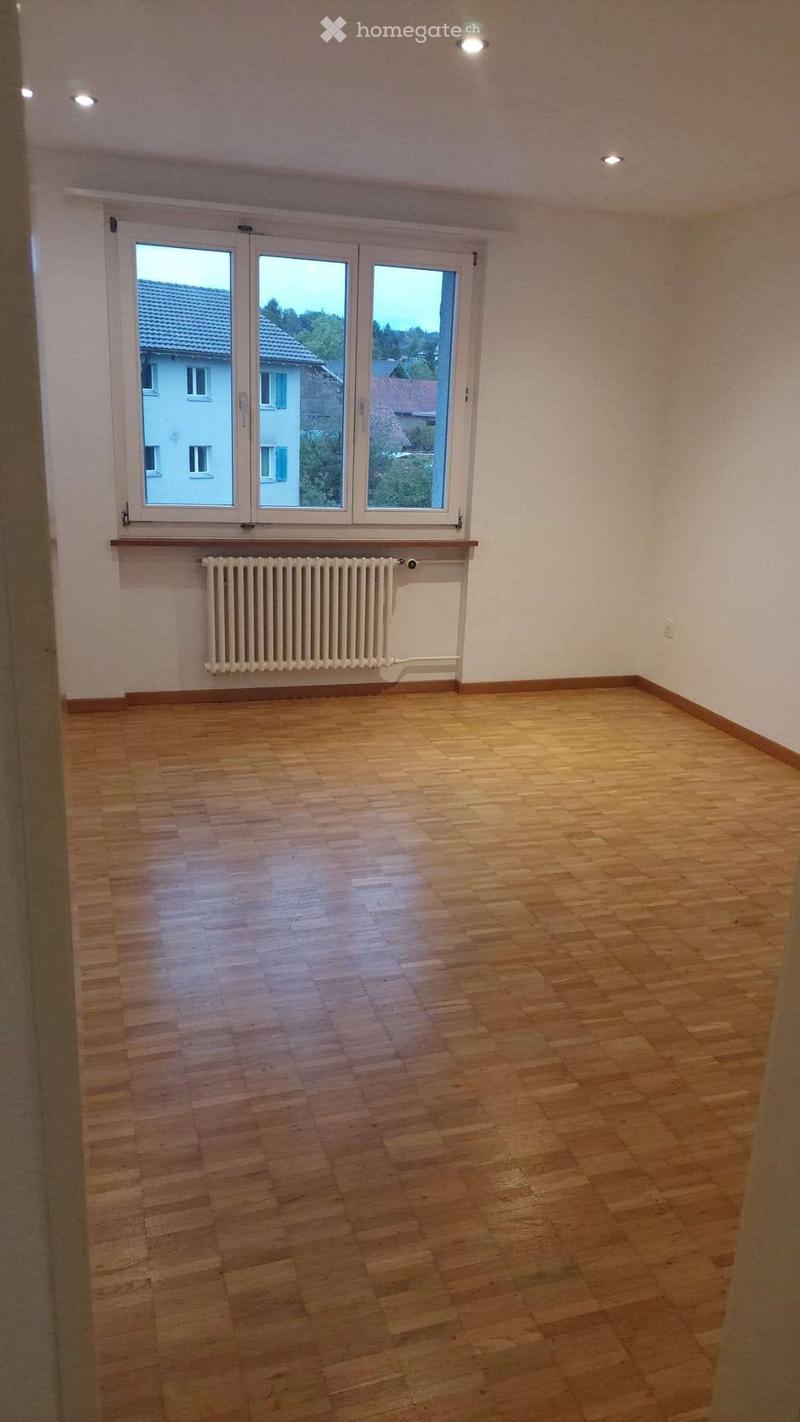 Empty room with wooden flooring, white walls, large windows, and a radiator below the windows.