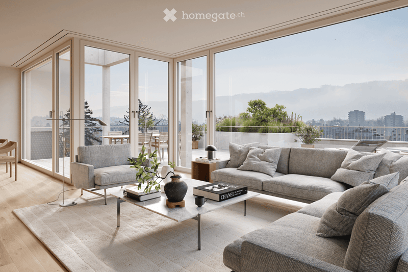 Modern living room with large windows offering a scenic view, featuring a grey sectional sofa, a coffee table with books and a vase, a floor lamp, and a dining area visible through the glass doors.
