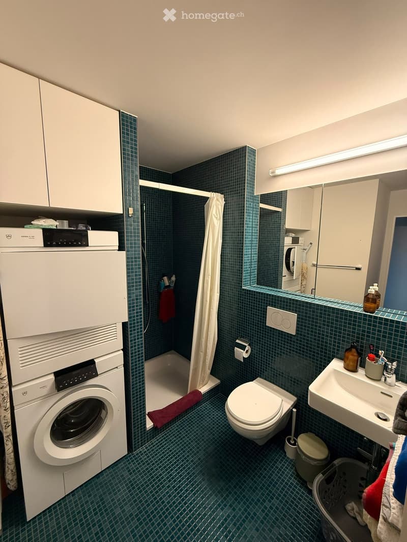 Compact bathroom with dark green tiled walls and floor, featuring a toilet, sink, mirror, shower, and a stacked washing machine/dryer unit.