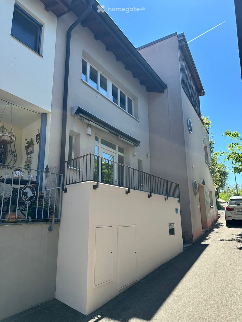 Two-story house with light beige exterior, multiple windows, and a balcony with metal railing. The house features a raised ground floor and a small garden area visible on the left side.