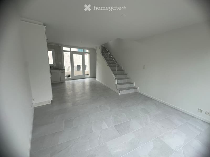 Empty room with light grey tiled flooring, white walls, and a staircase leading upwards. A small kitchen area is visible to the left with cabinets and a sink.