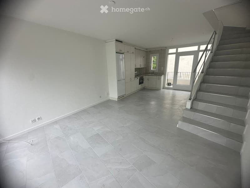 Empty room with grey tiled floor, white walls, staircase leading upwards, and a small kitchen area with cabinets and appliances.