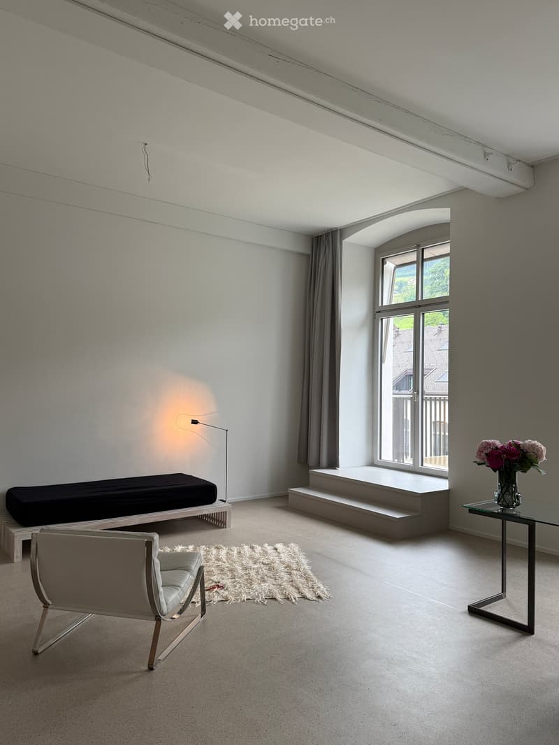 Modern, minimalist room with a large window, a bed, a chair, a glass table with a vase of flowers, a floor lamp, and a white shag rug.