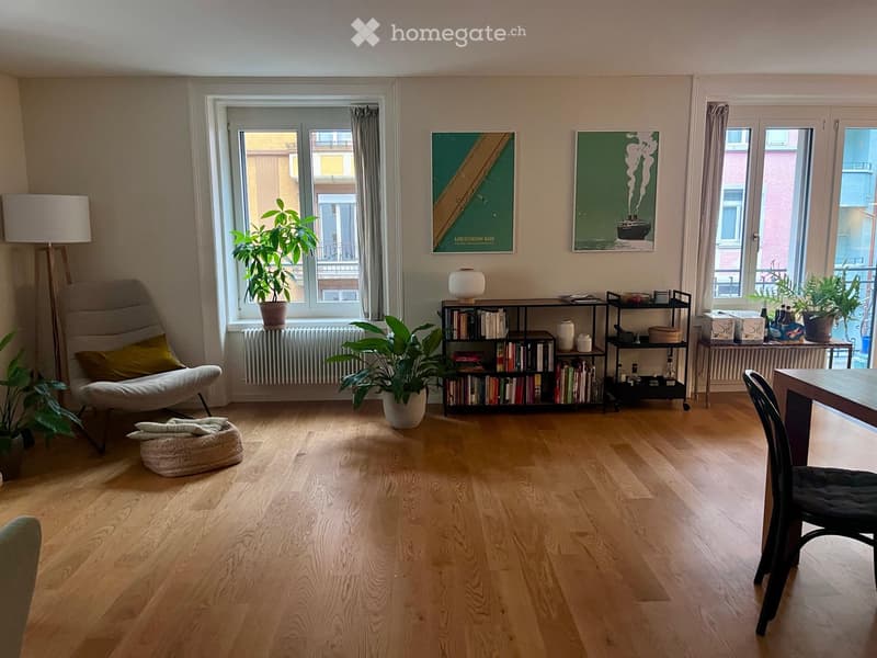 A modern living room with wooden flooring, large windows, a sofa, a chair, a table, a bookshelf, a lamp, and several potted plants.