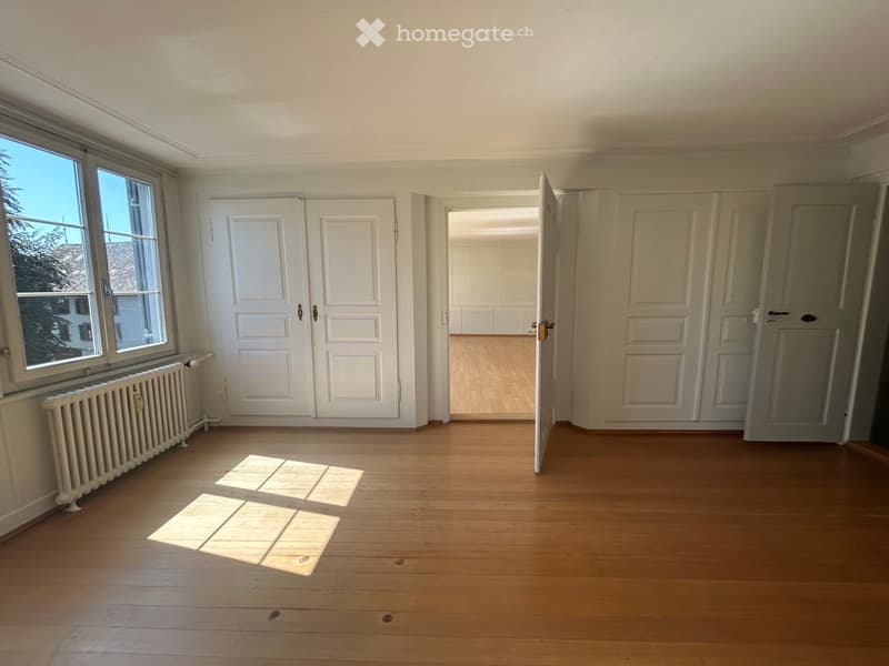 Empty room with wooden flooring, white walls, large windows, and several doors leading to other rooms.