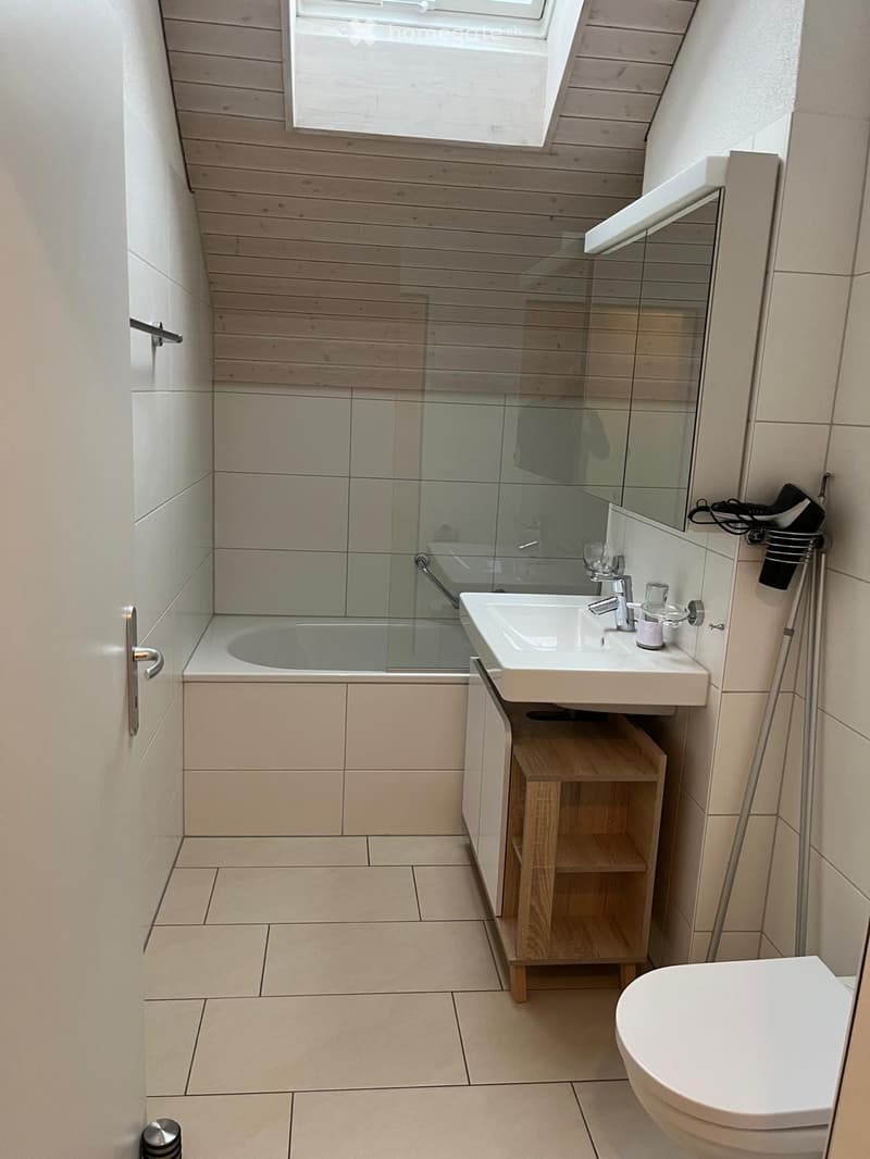 Modern bathroom with a white sink, bathtub, toilet, and a wooden shelf. Features a large mirror and a skylight window.