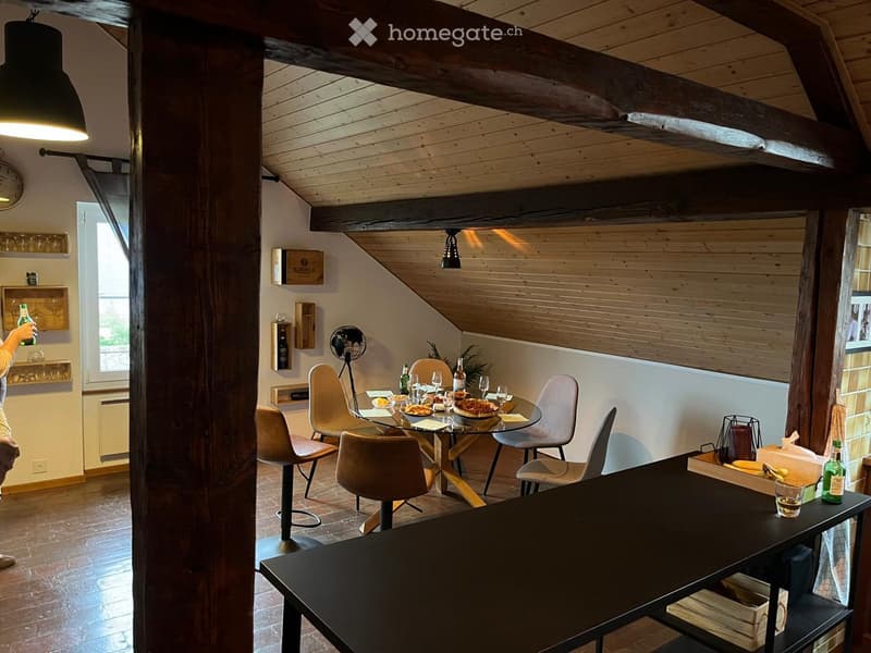 A modern, open-plan dining and lounge area with wooden beams, a round glass dining table surrounded by chairs, a bar counter, shelves, and a fan. The space features exposed wooden beams and warm lighting.