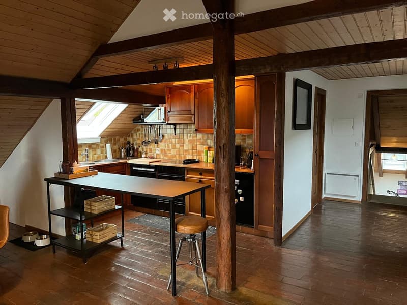The image shows a cozy kitchen with wooden beams and exposed wooden ceiling. The kitchen features wooden cabinets, a sink, a stove, and a refrigerator. There is a small table with a stool, a shelf with various items, and a cutting board on the counter. Th