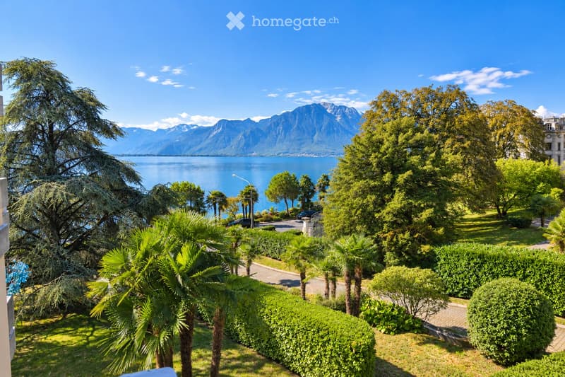 A lush garden with palm trees, neatly trimmed hedges, and a scenic view of a lake and mountains in the background.