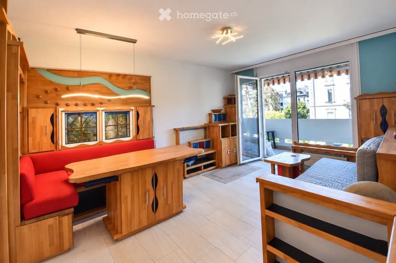 A modern, well-lit living space with a red sofa, wooden table, and a bed area, featuring built-in wooden cabinets and shelves.