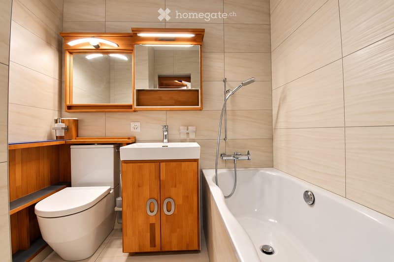 Modern bathroom with a white bathtub, toilet, sink, and wooden cabinet. Features a large mirror with built-in lighting above the sink.