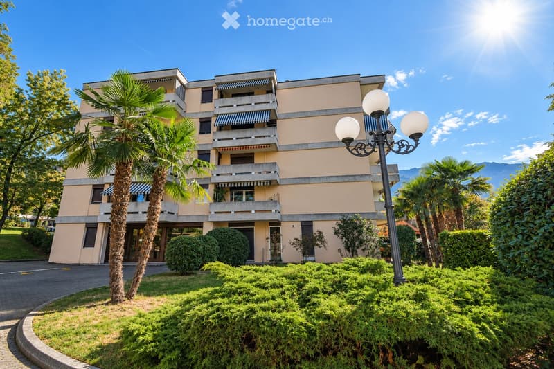 A multi-story residential building with beige walls, balconies, and striped awnings. The building is surrounded by well-maintained greenery, including palm trees and shrubs, with a decorative street lamp in the foreground.