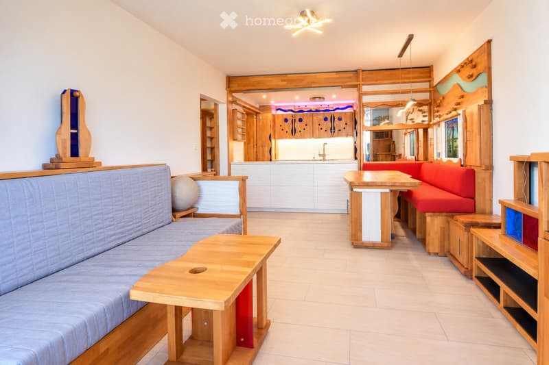 Modern living room with wooden furniture, including a blue sofa, wooden coffee table, red cushioned bench, and shelves. The room features a kitchen area in the background with white cabinets and a sink.