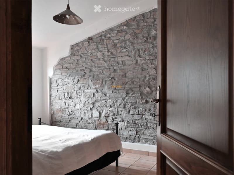 The image shows a bedroom with a stone accent wall, a bed with a white comforter, and a pendant light fixture. The room has a cozy and rustic feel with the exposed stone wall.