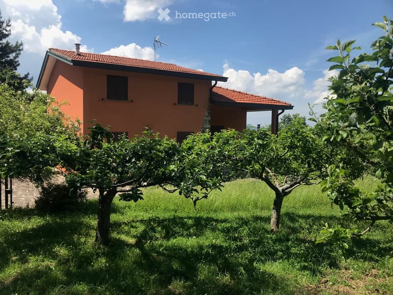 Two-story house with red tile roof, surrounded by lush greenery including fruit trees and a well-maintained lawn. The house has several windows and appears to have a balcony or terrace on the upper level.