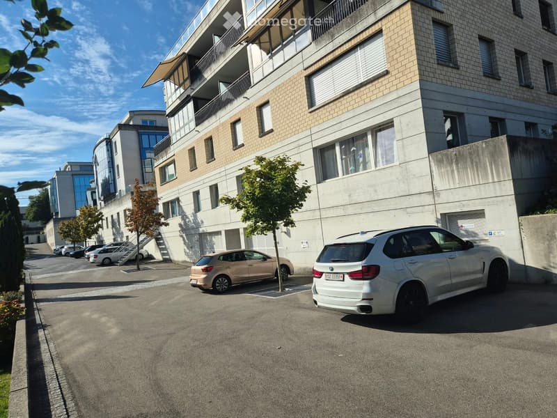 Modern multi-story building with balconies and large windows, surrounded by a parking area with several cars and small trees.