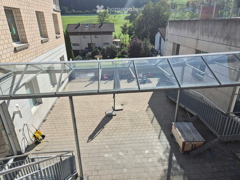 Balcony with a glass roof, tiled floor, metal railing, and a small wooden bench. The view shows a green field and a neighboring building.