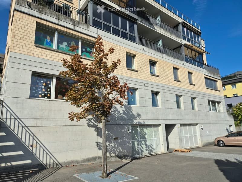 Four-story residential building with beige brick facade, large windows, balconies, and a tree in front.