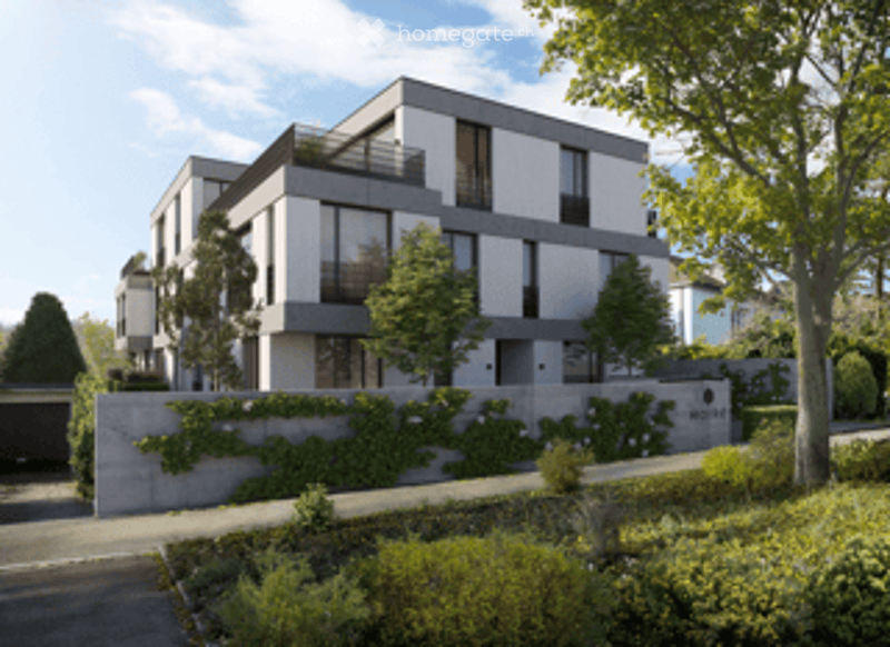Modern multi-story residential building with clean lines, large windows, and balconies. Features a garage entrance and landscaped surroundings with trees and shrubs.