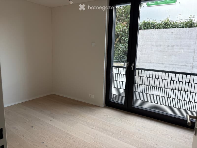 Empty room with light wooden flooring and beige walls, featuring a large sliding glass door leading to a balcony with a metal railing.