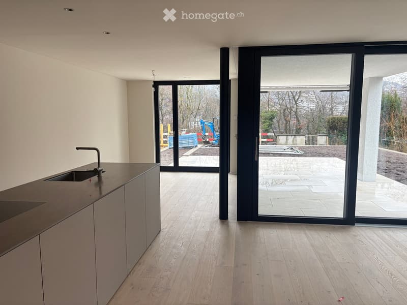 Empty kitchen with light wooden flooring, modern light-colored cabinets, and a sink with a faucet. Large sliding glass doors open to an outdoor area with construction equipment visible.
