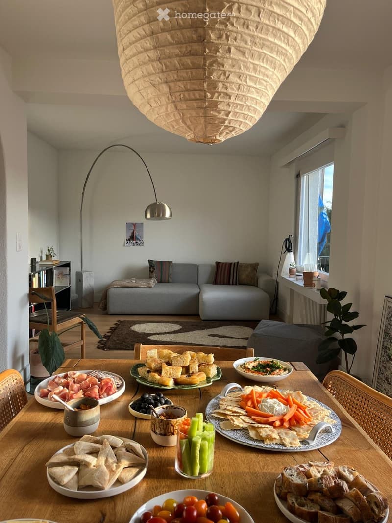 A cozy living room with a grey sofa, a wooden dining table set with various food items, a floor lamp, and a bookshelf in the background.