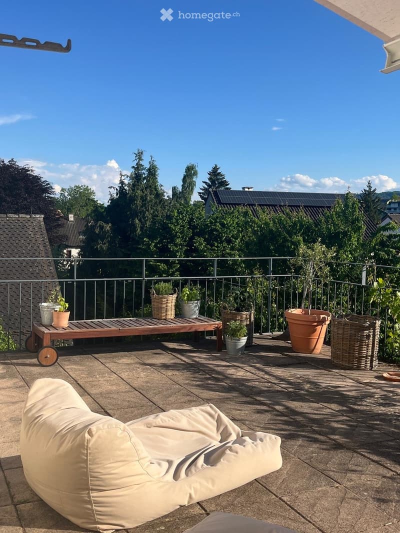 Balcony with a beige sofa, wooden table, potted plants, and a metal railing overlooking a green area.