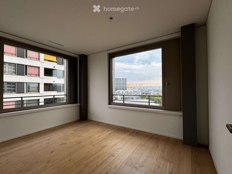 Empty room with wooden flooring, large windows offering a cityscape view, and neutral-colored walls.
