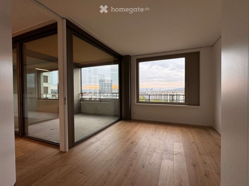 Empty room with wooden flooring, large glass sliding doors leading to a balcony, and a window offering a cityscape view.